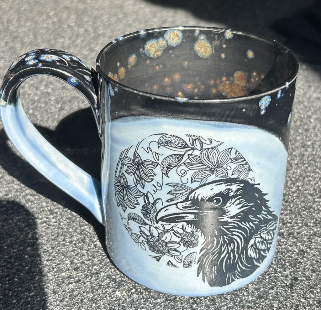 ceramic coffee mug in shades of blue with a black raven's head against a background of entwined leaves and flowers