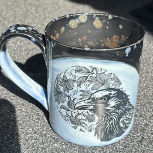 ceramic coffee mug in shades of blue with a black raven's head against a background of entwined leaves and flowers